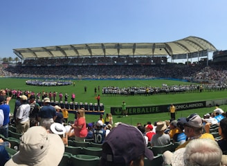 A large sports stadium filled with spectators, a well-maintained green field in the center, and a group of people forming lines as they prepare for an event. The seating areas are crowded, and the atmosphere appears lively with fans wearing various team colors and merchandise. Overhead, clear skies and bright sunlight illuminate the venue.