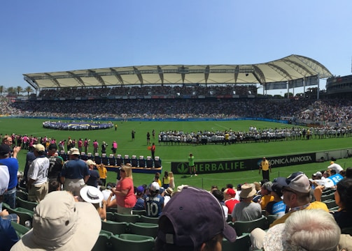 A large sports stadium filled with spectators, a well-maintained green field in the center, and a group of people forming lines as they prepare for an event. The seating areas are crowded, and the atmosphere appears lively with fans wearing various team colors and merchandise. Overhead, clear skies and bright sunlight illuminate the venue.