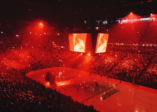 An indoor ice hockey arena filled with a large crowd, bathed in red lighting. A giant central scoreboard projects fiery visuals, illuminating the ice and audience. The atmosphere is charged, likely during a live sporting event.
