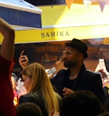 A lively crowd gathers in an outdoor setting with many people capturing the moment on their phones. The atmosphere is cheerful and energetic, with some individuals wearing sports jerseys, likely celebrating or supporting an event.