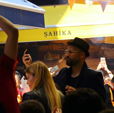 A lively crowd gathers in an outdoor setting with many people capturing the moment on their phones. The atmosphere is cheerful and energetic, with some individuals wearing sports jerseys, likely celebrating or supporting an event.