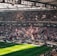 A packed sports stadium filled with enthusiastic fans waving flags and banners. The crowd is dense, showcasing a dynamic and lively atmosphere. Sunlight filters through the stadium roof, casting patterns on the green field below.