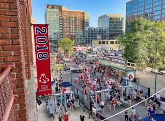 A vibrant outdoor scene featuring a large crowd of people gathered near a sports venue. A prominent red banner with '2018' and a sports logo hangs from a brick building. Nearby, there is a sign for a team store and souvenir shop. Surrounding the event are several modern high-rise buildings and a parking lot full of cars. Tall trees are visible in the background.