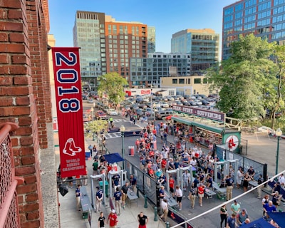 A vibrant outdoor scene featuring a large crowd of people gathered near a sports venue. A prominent red banner with '2018' and a sports logo hangs from a brick building. Nearby, there is a sign for a team store and souvenir shop. Surrounding the event are several modern high-rise buildings and a parking lot full of cars. Tall trees are visible in the background.
