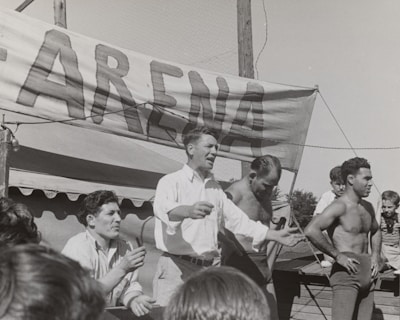 A group of men, some shirtless, are gathered around a platform or stage. A man in a white shirt appears to be speaking or gesturing to a crowd. A large sign with the word 'ARENA' is prominently displayed in the background. The scene seems to be set outdoors on a sunny day.
