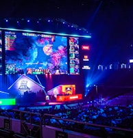 A large esports arena featuring a massive screen displaying a video game, surrounded by sponsor logos and team names. The auditorium is filled with spectators watching the gaming event. Neon blue and purple lights illuminate the stage, creating a vibrant atmosphere.