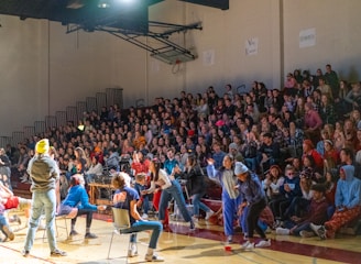 A crowded gymnasium with many people seated on bleachers watching an event. Several individuals are actively participating on the gym floor, some of whom are wearing casual clothing and engaging in activities. The atmosphere appears lively and energetic.