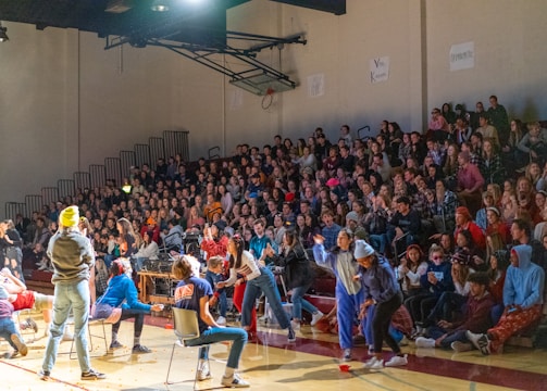 A crowded gymnasium with many people seated on bleachers watching an event. Several individuals are actively participating on the gym floor, some of whom are wearing casual clothing and engaging in activities. The atmosphere appears lively and energetic.