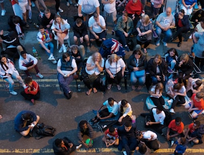 A large group of people gathered outdoors, watching a screen that is displaying a man. The crowd is diverse in age and appears interested and engaged, with many sitting closely together, some standing. Many are dressed casually, some in sports attire, and a few waving or holding flags. The event seems to have a communal, festive atmosphere.
