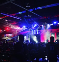A dimly lit indoor arena filled with an audience. Bright stage lights in red and blue illuminate multiple large screens displaying graphics.