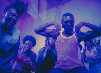 A group of people appear to be enjoying themselves in a lively, colorful environment. A man in the center strikes a pose with his arms flexed, wearing sunglasses and a mesh tank top, while others around him are smiling and cheering. The lighting casts a purple and blue hue over the scene, adding to the energetic and dynamic atmosphere.