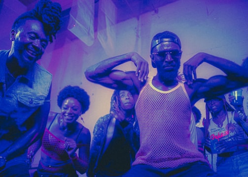 A group of people appear to be enjoying themselves in a lively, colorful environment. A man in the center strikes a pose with his arms flexed, wearing sunglasses and a mesh tank top, while others around him are smiling and cheering. The lighting casts a purple and blue hue over the scene, adding to the energetic and dynamic atmosphere.