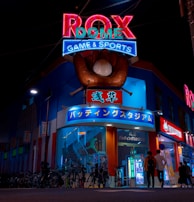 A vibrant, neon-lit facade of a gaming and sports establishment at night, featuring a large glove holding a baseball above the entrance. The building is adorned with colorful signs in both English and Japanese, with various advertisements and an illuminated sign that says 'ROX DOME'. Several people are walking towards the entrance, and bicycles are parked along the side.