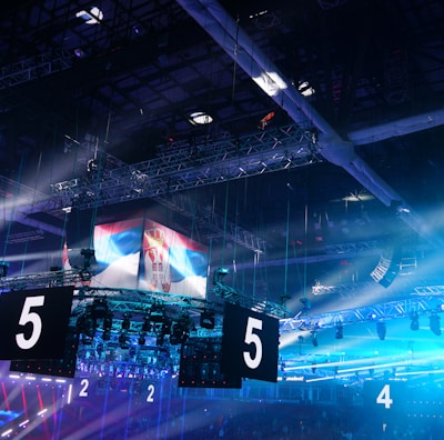A large indoor arena is equipped with advanced lighting and audio systems. Suspended above the space are numerous digital displays showcasing numbers. The environment is filled with blue illumination and spotlights creating a vibrant atmosphere.