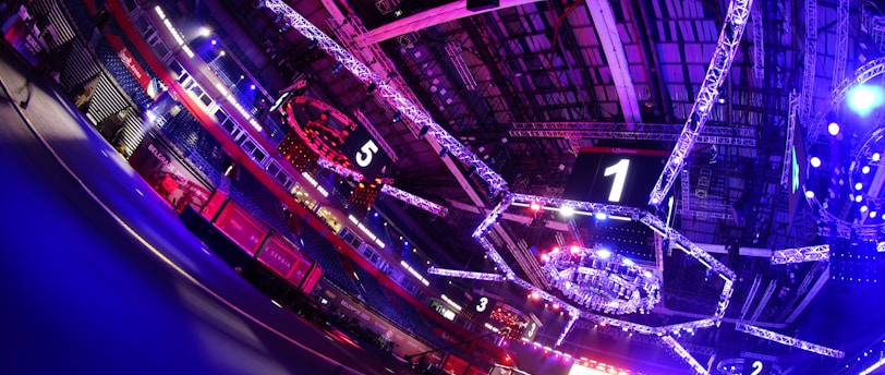 An indoor arena with vibrant purple and blue lighting, featuring large numbered screens suspended from the ceiling. The venue has a high ceiling with intricate metal truss structures and hanging lights, creating a futuristic and energetic atmosphere.