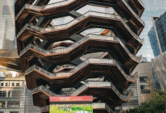 A large, spiral architectural structure with multiple levels stands prominently in an urban setting. In front of the structure, a large screen displays a sports broadcast, and several people are gathered around, some sitting and others standing. The scene is set outdoors, with towering modern buildings surrounding the area. The ground is paved with a geometric pattern of gray and white tiles.