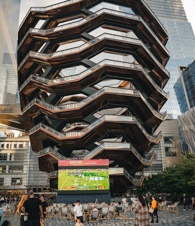 A large, spiral architectural structure with multiple levels stands prominently in an urban setting. In front of the structure, a large screen displays a sports broadcast, and several people are gathered around, some sitting and others standing. The scene is set outdoors, with towering modern buildings surrounding the area. The ground is paved with a geometric pattern of gray and white tiles.